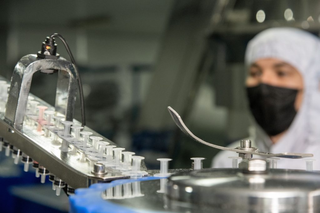 Close-up of a pharmaceutical production line with plastic syringe components moving along a conveyor, while a technician in cleanroom attire monitors the process in the background.