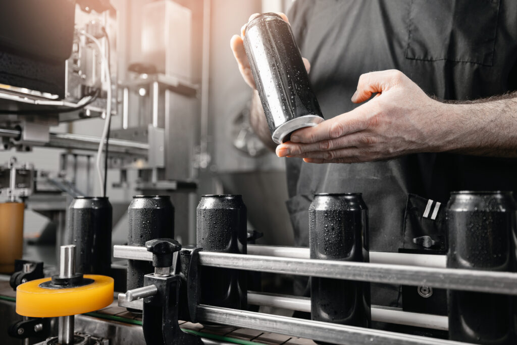 Maintenance technician inspecting black beverage cans on a production line during top load quality control testing in a beverage manufacturing facility.